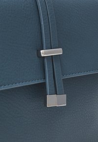 Sac en cuir bleu à surface texturée, doté de ferrures argentées et de deux fines bretelles fixées par des motifs métalliques carrés.