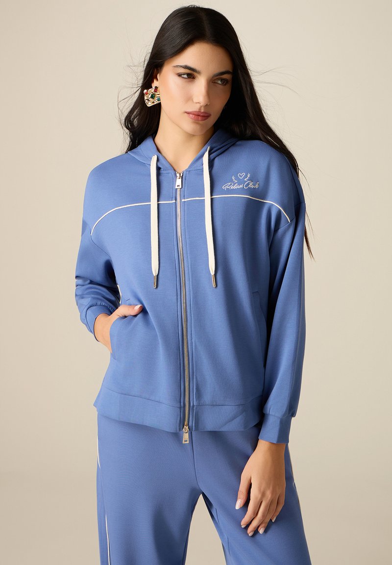 Mujer con sudadera azul con cremallera y cordones blancos, y pantalones a juego, de pie con una mano en el bolsillo contra un fondo liso.