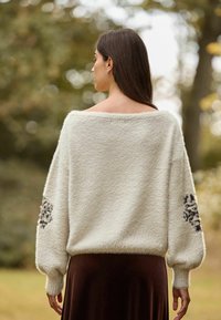 Weißer, flauschiger, übergroßer Pullover mit schulterfreiem Design, strukturiertem Stoff und schwarzer Blumenstickerei an den Ärmeln. Dunkelbrauer Rock ist darunter sichtbar.
