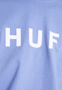 Světle modré bavlněné tričko s výraznými bílými blokovými písmeny, které hlásají "HUF". Hladká textura, design s kulatým výstřihem. Minimalistický styl.