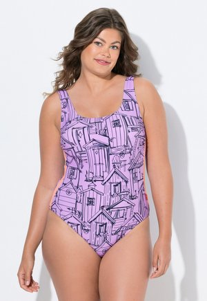 Kvinde med bølget brunt hår iført en lilla heldragt badedragt med sorte skitser af små huse, stående mod en hvid baggrund.
