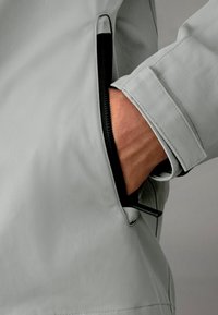 Jachetă din țesătură gri deschis, cu un buzunar cu fermoar cu margine neagră, textură netedă și linii curate, evidențiind un design modern.