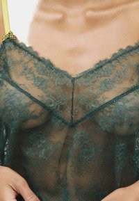 Zielony koronkowy top z kwiatowym wzorem, dekoltem w serek, przejrzystym materiałem i żółtymi ramiączkami z kokardką.