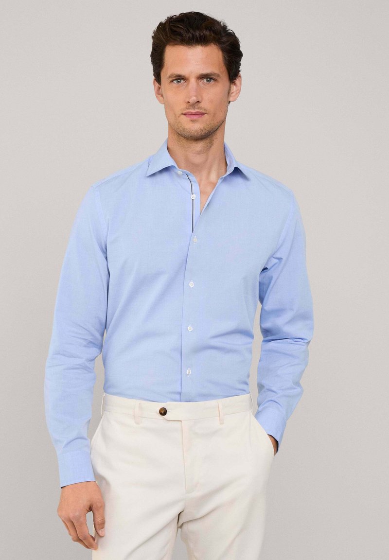 Camisa de algodón azul claro con mangas largas, frente abotonado y cuello puntiagudo, combinada con pantalones color crema.