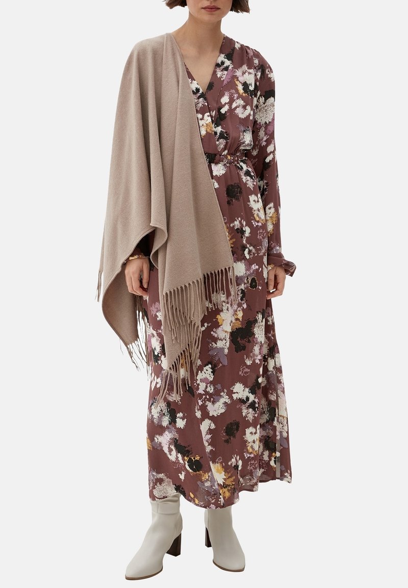 Femme portant une longue robe fleurie mauve avec un châle beige drapé sur une épaule et des bottines blanches.