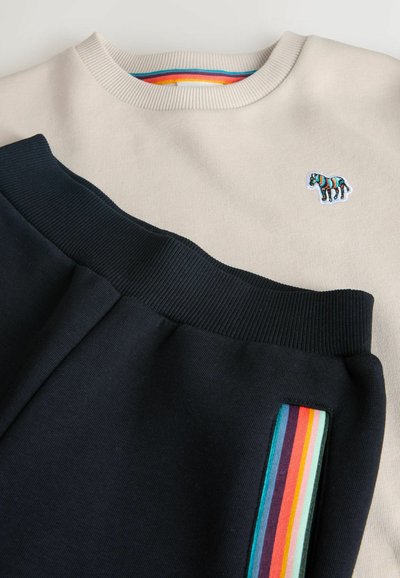 Béžová mikina s rebrovaným golierom a malým multicolor zebrovým logom, v kombinácii s námorníckymi joggermi so zvýraznením v dúhových prúžkoch.