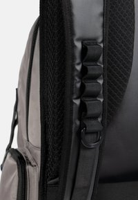 Rucksack mit grauem Stoff, einer strukturierten schwarzen Mesh-Panel, verstellbarem Riemen und zwei seitlichen Reißverschlussfächern.