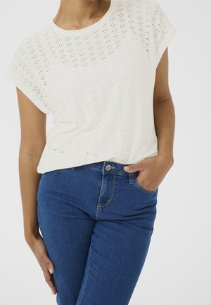 Femme portant un haut blanc à manches courtes en tissu ajouré et un jean bleu taille haute, une main dans une poche et l'autre reposant sur sa cuisse.