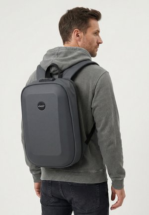 Mężczyzna w szarej bluzie z kapturem i czarnych dżinsach niesie na plecach szary plecak Venezia o twardej konstrukcji, stoi bokiem.