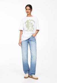 Weißes oversized T-Shirt mit grünem botanischen Grafikdesign und Schriftzug, kombiniert mit hellblauen Straight-Leg-Jeans und beigen Sneakers.
