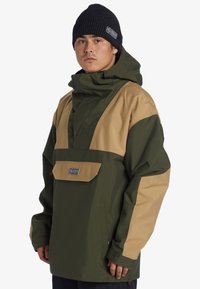 Chaqueta impermeable verde y marrón con capucha, que cuenta con un bolsillo frontal, paneles de color contrastante y una etiqueta de marca en el bolsillo.