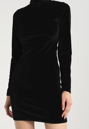 Fitted black velvet long-sleeve mini dress with high neckline, worn by person standing against plain background.