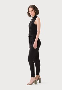 Černý overal s halter krkem, bočním vázáním v pase a zúženými nohavicemi. Modelka má na sobě zelené sandály na blokovém podpatku.