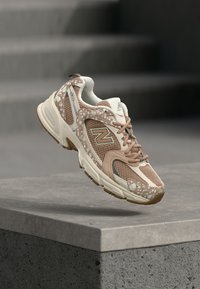 Αθλητικό παπούτσι New Balance σε μπεζ και καφέ χρώμα με δίχτυ και διακοσμητικά σχέδια να αιωρείται πάνω από μια γκρι τσιμεντένια επιφάνεια.