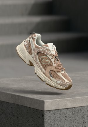 Beige and brown New Balance sneaker with mesh and patterned overlays hovering above a gray concrete ledge.