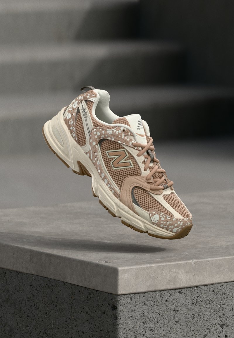 Αθλητικό παπούτσι New Balance σε μπεζ και καφέ χρώμα με δίχτυ και διακοσμητικά σχέδια να αιωρείται πάνω από μια γκρι τσιμεντένια επιφάνεια.