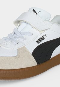 Λευκό sneaker Puma με μαύρο λογότυπο, μπεζ σουέντ λεπτομέρειες, λευκά κορδόνια, λουράκι με βέλκρο και καφέ ελαστική σόλα.