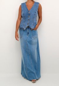 Frau, die eine ärmellose Jeansweste mit Knöpfen und einen langen Jeansrock mit vorderen Taschen und rohem Saum trägt, steht mit einer Hand in der Tasche.