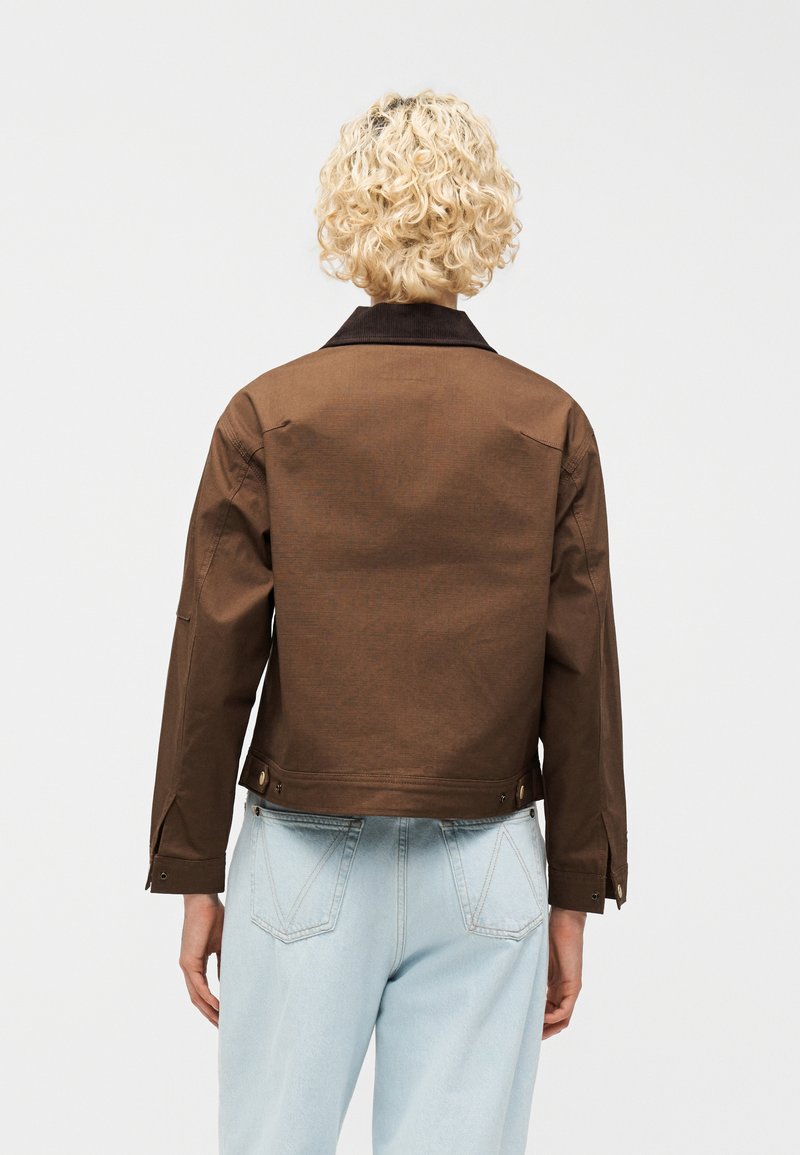 Persona bionda con capelli ricci che indossa una giacca marrone con colletto nero e jeans azzurri, mostrata di spalle su uno sfondo semplice.