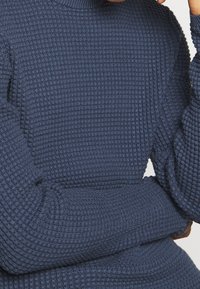 Gros plan d'une personne portant un pull en tricot texturé bleu foncé à manches longues, les bras croisés et une main touchant le cou.