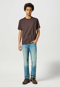 T-shirt di cotone marrone a maniche corte, abbinato a jeans di denim azzurri. Scarpe in suede marroni con suole in gomma nera completano l'outfit.