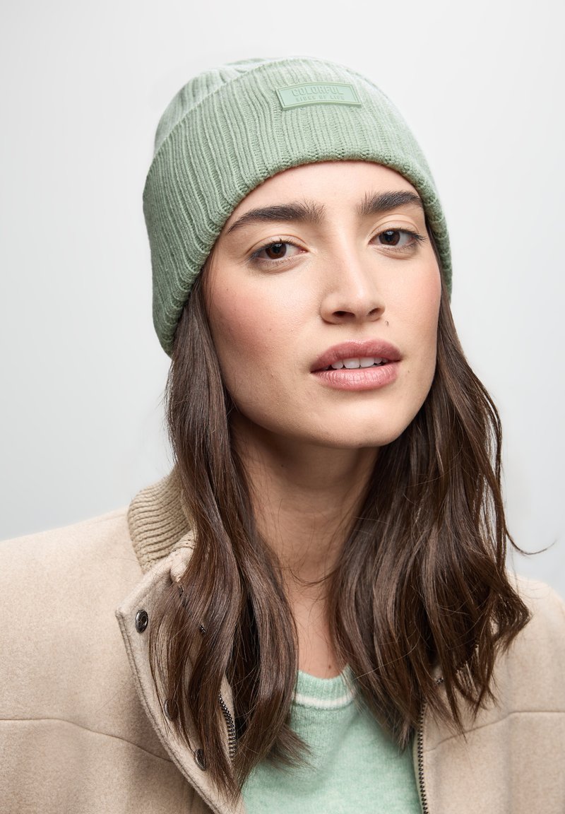 Berretto a coste in maglia verde con una toppa con logo, indossato con una giacca beige. Il tessuto sembra morbido e il design è aderente, coprendo la testa.