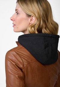 Bruine leren jas met een zwarte capuchon eraan bevestigd. Gladde textuur, slim-fit ontwerp. De jas heeft gedetailleerd stiksel langs de schouders.