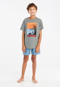 Camiseta de algodón gris con un gráfico colorido que dice "Siente la Ola", combinada con pantalones cortos azules con pequeños patrones de palmeras.