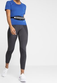 Camiseta deportiva azul real, bolsa negra con cintura, y leggings negros con rayas. Zapatillas blancas con acentos morados. Detalles de tela texturizada.