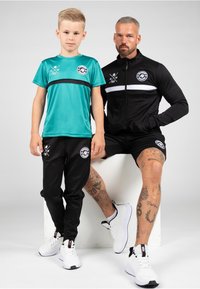 Child in a teal sports shirt with black accents and black pants. Adult in a black zip-up jacket and shorts. Both wear white sneakers.