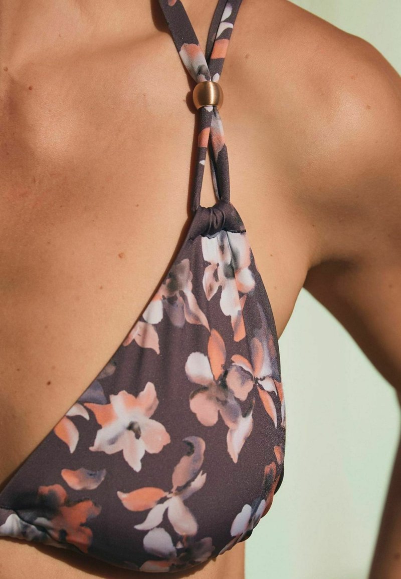 Bikini top s kvetinovým vzorom v ružovej, bielej a šedej farbe, ktorý nosí osoba s opálenou pokožkou na svetlozelenom pozadí.