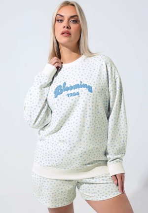 Donna con lunghi capelli biondi che indossa una felpa bianca e pantaloncini abbinati con un piccolo motivo floreale blu e la scritta "Blooming 1986".