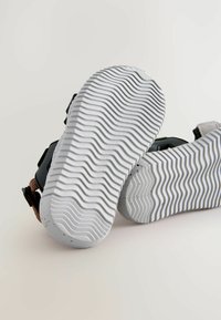 Melni sandales ar teksturētām pelēkām gumijas zolēm, kas veidotas viļņveidīgā rakstā. Regulējamas siksnas brūnā un pelēkā materiālā ar drošu sprādzi.