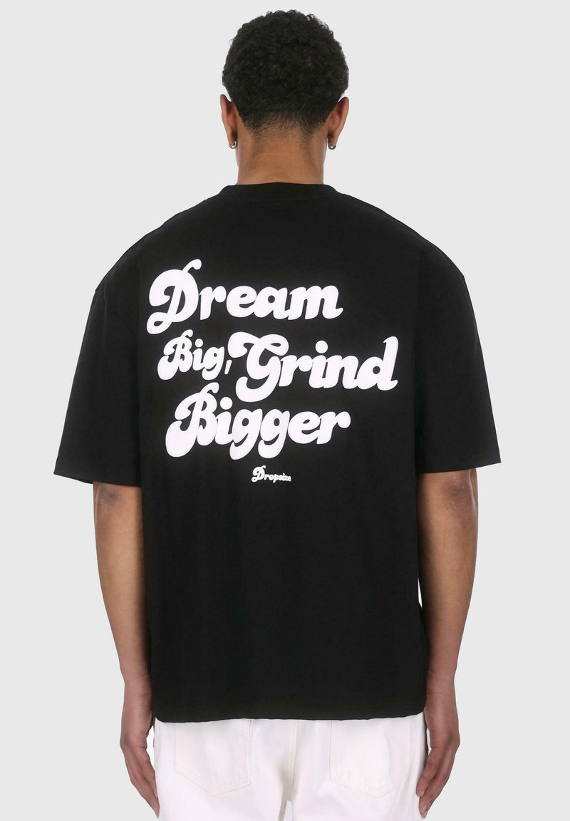 Schwarzes Baumwoll-T-Shirt mit einem großen weißen Textslogan auf der Rückseite: "Dream Big, Grind Bigger". Es hat eine entspannte Passform.