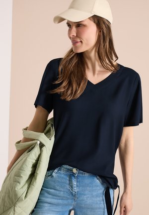 Femme portant une casquette beige, un t-shirt bleu marine à col en V, un jean bleu clair, tenant une veste matelassée vert clair sur un bras, regardant sur le côté.