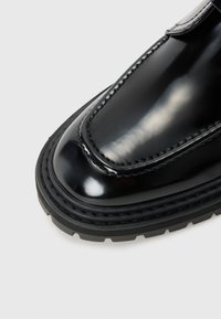 Zapato de charol negro con punta redondeada, costuras visibles y una suela de goma negra gruesa y texturizada. Diseño sencillo sin acentos adicionales.