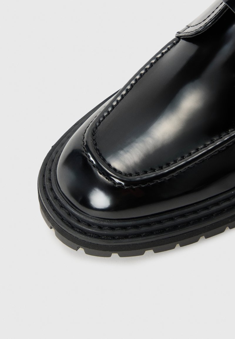 Scarpa in vernice nera con punta tondeggiante, cuciture visibili e una suola in gomma nera spessa e strutturata. Design semplice senza ulteriori decorazioni.