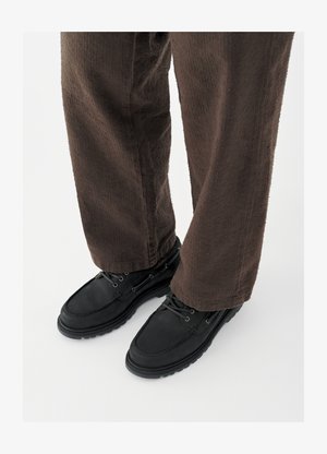 Svarte skinnsko med glatt finish, avrundet tå og snøresystem, kombinert med teksturerte brune bukser. Sett fra bakkenivå.