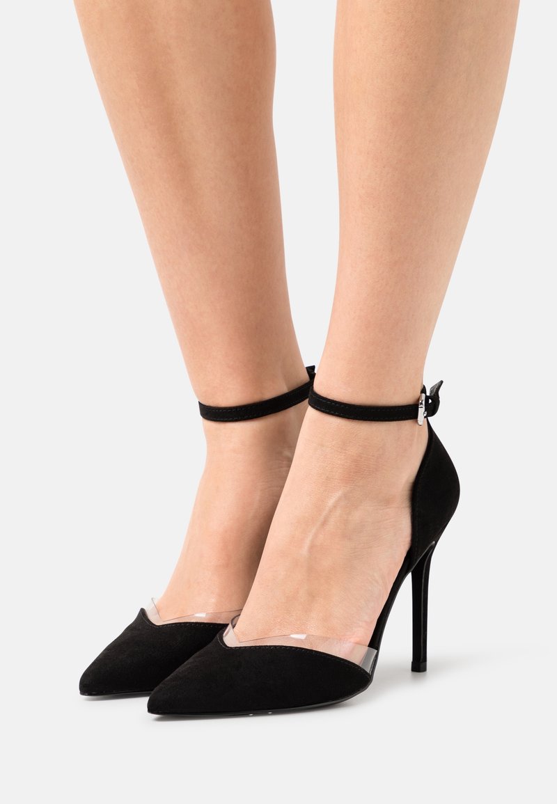 Jambes portant des escarpins noirs en daim à bouts pointus, avec des brides transparentes sur le dessus et des sangles fines à la cheville avec des boucles argentées.