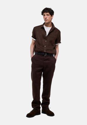 Homme aux cheveux bouclés debout, les mains dans les poches, portant une chemise marron sur un tee-shirt blanc rayé, un pantalon marron foncé, une ceinture noire et des chaussures marron.