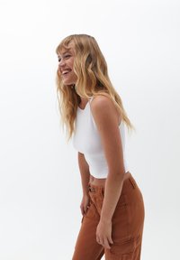 Top corto bianco a costine, pantaloni cargo arancioni con tasche laterali, vestibilità strutturata. Capelli acconciati in onde morbide, su uno sfondo bianco semplice.
