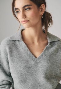 Maglione grigio a maglia con scollo a V e colletto. Texture morbida, dettagli a coste sul colletto. Modella con capelli castani di lunghezza media e orecchini a cerchio.