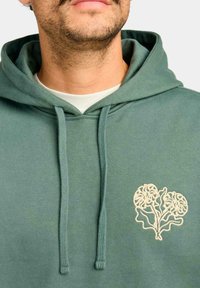Sudadera de algodón verde con dos cordones, que presenta un bordado floral en forma de corazón beige en el área del pecho izquierdo. Textura suave.