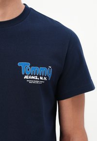 Tamsiai mėlyna marškinėlė su "Tommy Jeans, N.Y." logotipu mėlynos ir baltos spalvos kairėje krūtinės pusėje, dėvima asmens su šviesiai rudais odos atspalviais.
