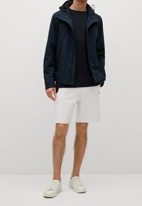 Tmavě modrá voděodolná bunda s kapucí, černé tričko pod ní, bílé kraťasy a bílé tenisky. Hladká látka a zapínání na knoflíky vepředu.