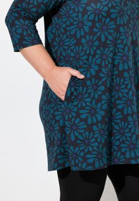 Sininen kukallinen tunika, jossa on kolme neljäsosaa hihat, tummalla taustalla ja sivutaskuilla, valmistettu pehmeästä ja joustavasta kankaasta.