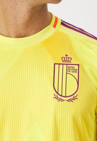 Yellow athletic jersey featuring a purple crest with "ROYAL BELGIAN FA 1895," vertical stripe pattern, and ribbed collar with red and purple accents.