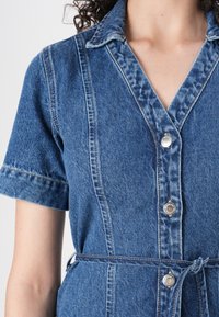 Denim obleka s kratkimi rokavi, ovratnikom in gumbom spredaj. Ima pas z vezalno vrvico in subtilne šive, v srednje modri barvi.