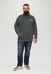 Grijze sweater met hoge kraag, voorzak en vervaagde textuur, gecombineerd met donkerblauwe jeans en beige sneakers met zwarte accenten.