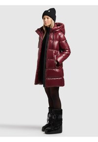 Mujer con un abrigo acolchado brillante color granate con capucha, gorro negro, vestido negro, medias transparentes y botas negras de invierno, de perfil.